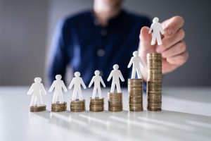 human hand placing small human figure on increasing stacked coins over wooden desk