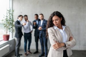 Workplace Sexism And Bullying, Unhappy Victimized Businesswoman Standing While Her Male Colleagues Whispering Behind Her Back Standing In Modern Office